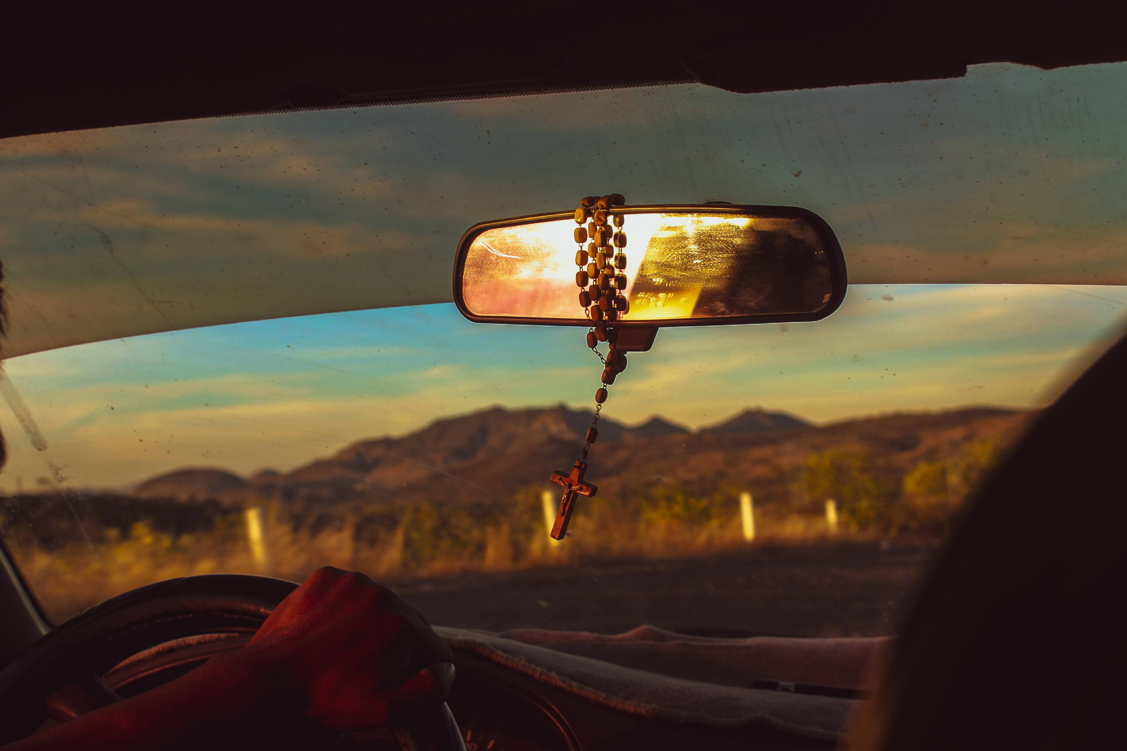 View through a car windshield at sunset with a rosary hanging from the rearview mirror and mountains in the distance